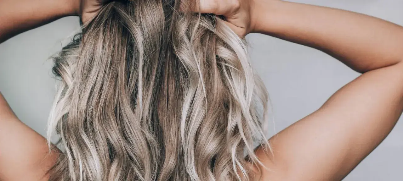 Frau mit blonden, welligen Haaren, die ihre Hände im Nacken hat. Sie zeigt, wie man die Haare pflegt, um sie gesund und glänzend zu halten.
