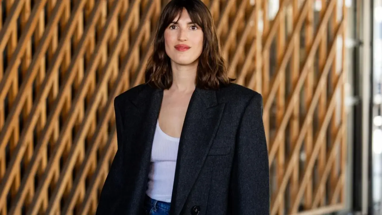 Frau mit französischem Crop-Haarschnitt, trägt einen Oversized-Blazer und Jeans vor einer Holzwand.