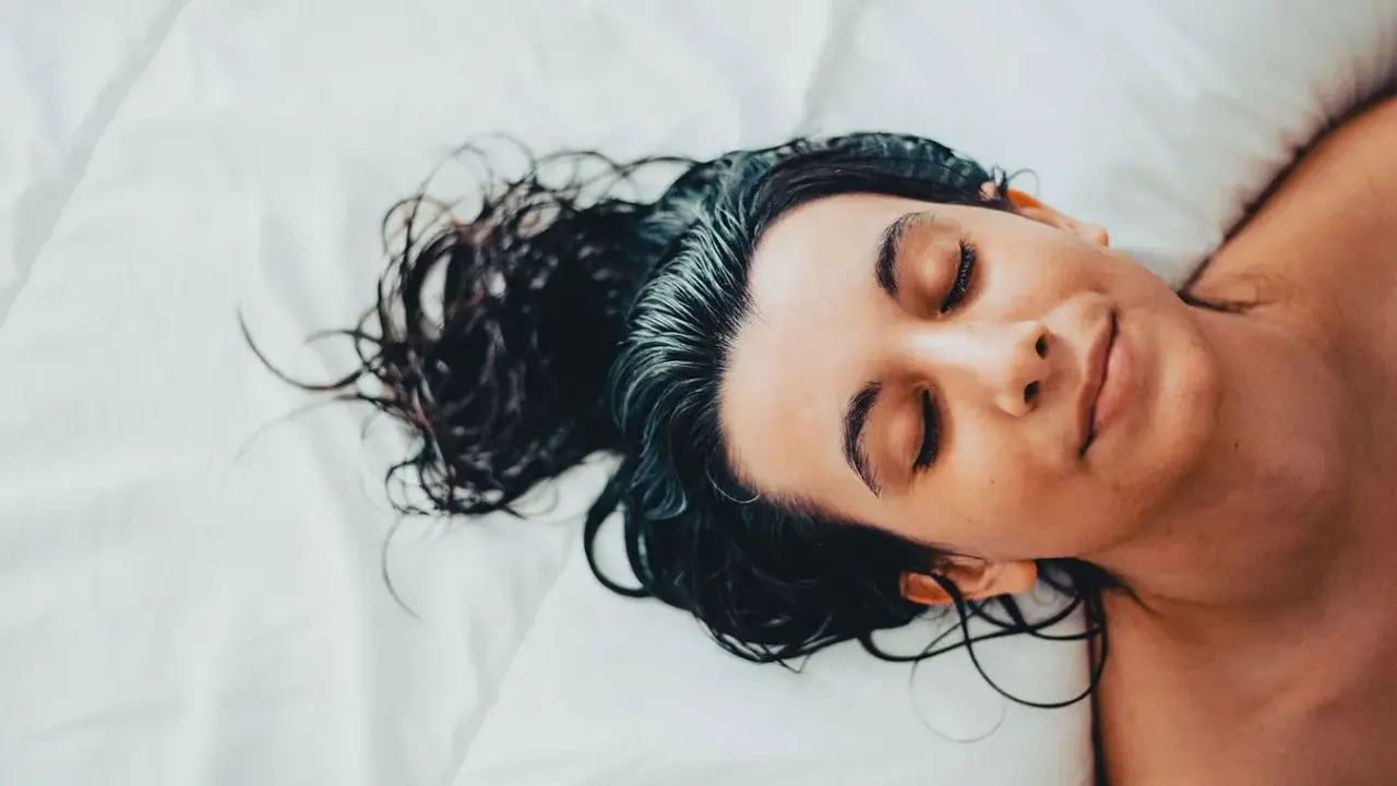 Eine Frau mit dunklen, nassen Haaren schläft auf einem weißen Bett. Sie liegt auf dem Rücken, die Augen geschlossen, und ihr Gesicht ist entspannt.