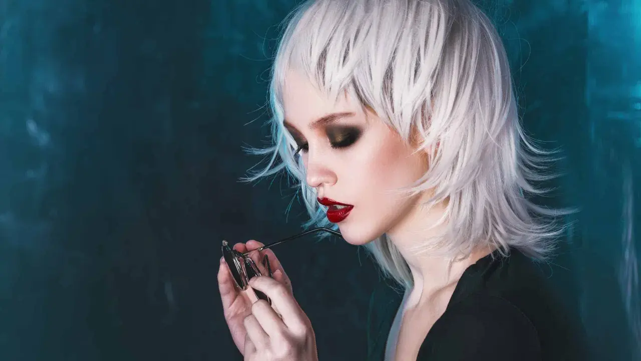 Stylische Dame mit weißem Wolfcut, dunklem Lidschatten und roten Lippen hält eine Brille.