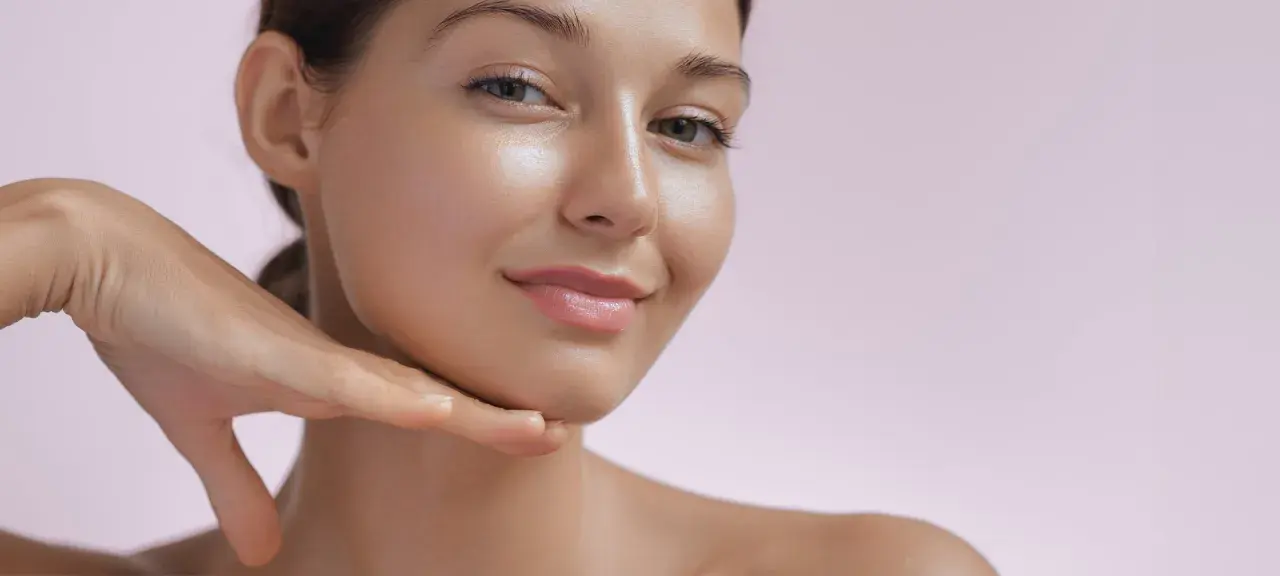 Ein Gesicht mit strahlendem, glowy Make-up und einem sanften Lächeln, die Hand berührt das Kinn.