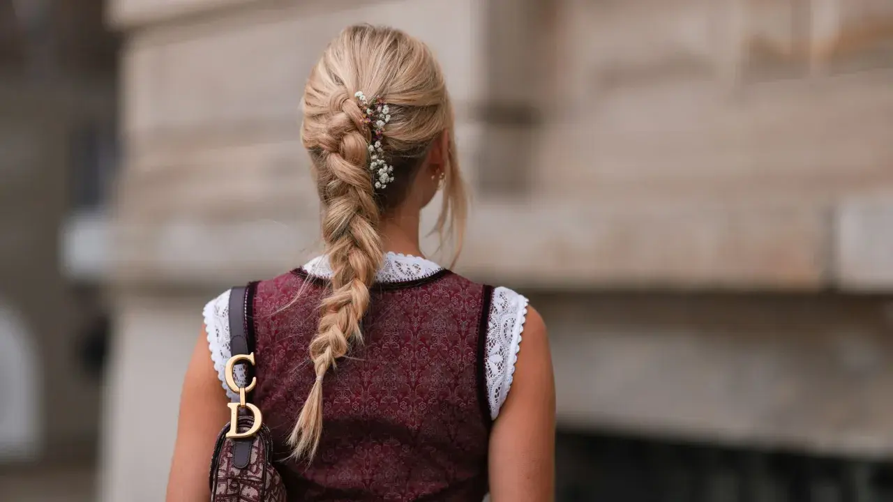 Blonde Zopf-Frisur mit Blumen-Akzenten, perfekt für Oktoberfest-Frisuren. Ein Dirndl und eine Dior-Tasche runden den Look ab.