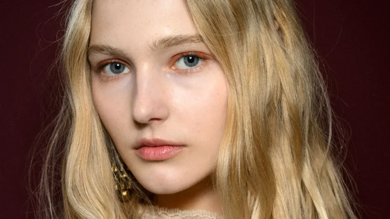 Blonde Haare umrahmen ein Gesicht mit blauen Augen und dezentem Make-up. Ihre hooded eyes wirken geheimnisvoll.