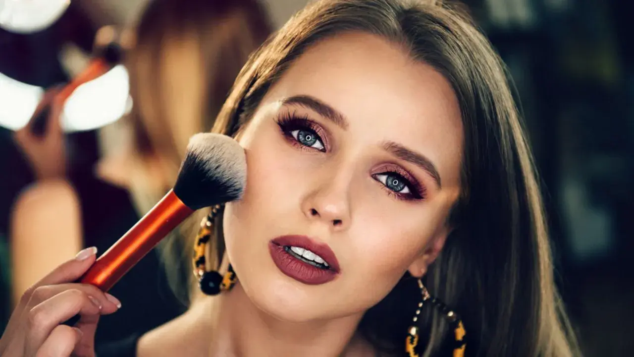 Ein glamouröses Abend Make-up wird aufgetragen. Eine Hand hält einen Pinsel, der über die Wange einer Frau mit dramatischem Lidschatten und Lippenstift gleitet.