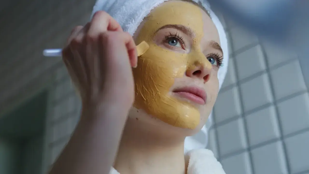 Junge Frau mit Handtuch auf dem Kopf trägt eine Gesichtsmaske auf. Die Kurkuma Wirkung auf die Haut ist bekannt für ihre entzündungshemmenden Eigenschaften.