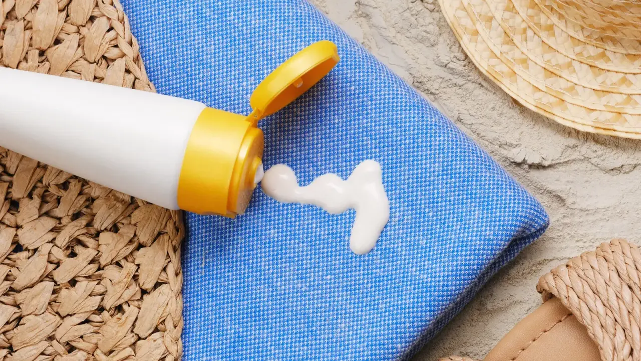 Sonnencreme-Flecken auf dem blauen Handtuch. Ein weißer Klecks Sonnencreme tropft aus einer Tube. Strandutensilien wie Hut und Sandalen liegen daneben.