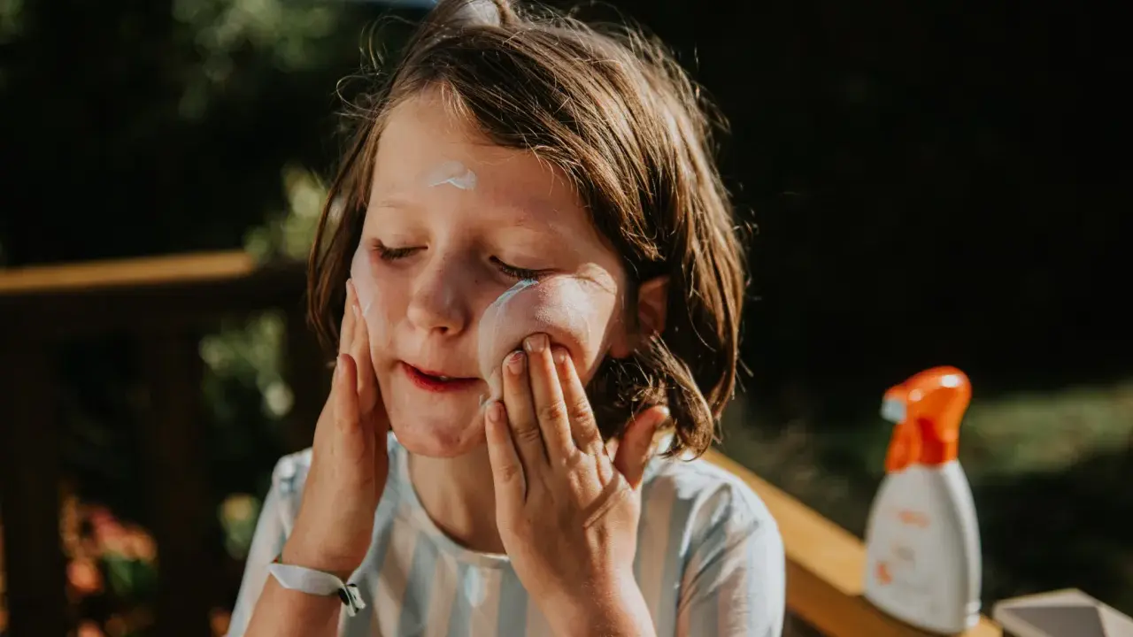 Kind mit geschlossenen Augen trägt Sonnencreme auf. Eine Flasche abgelaufene Sonnencreme steht daneben.