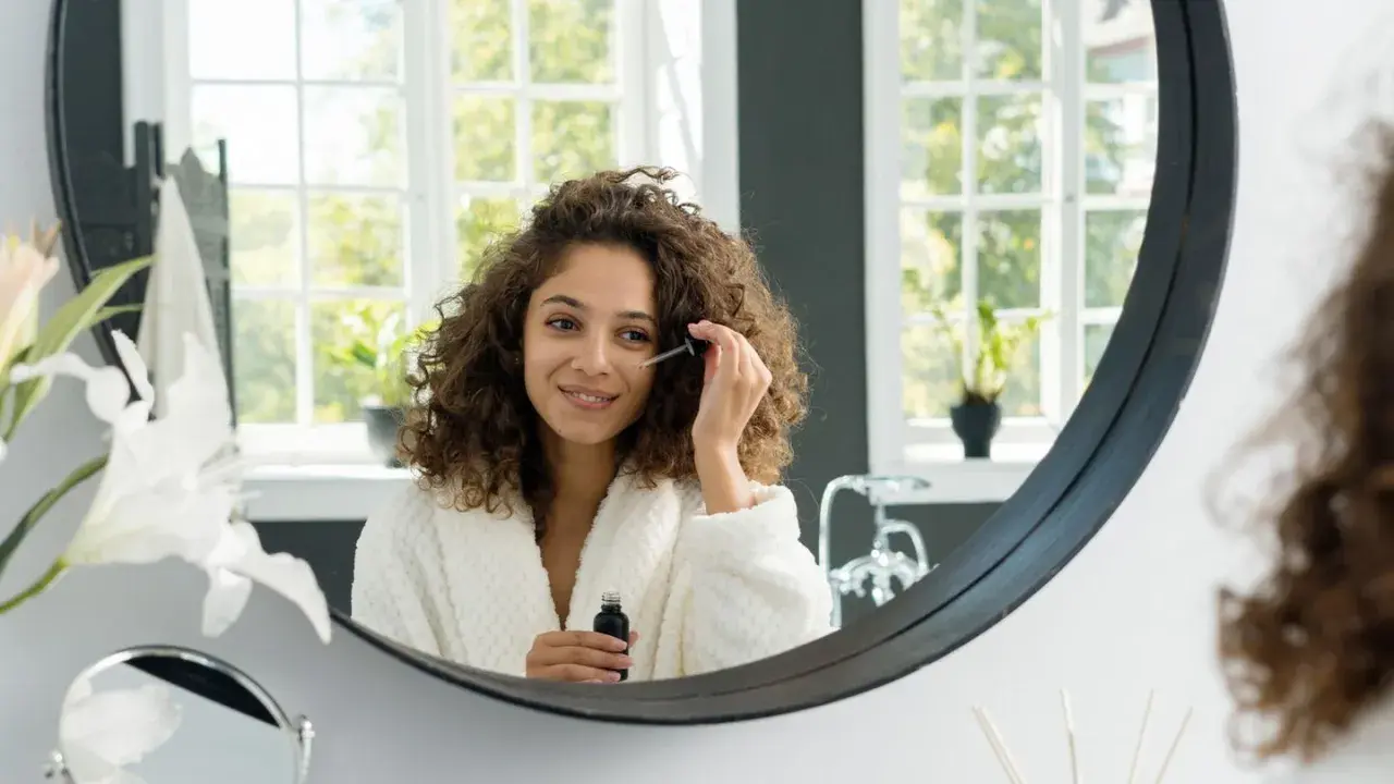 Frau mit lockigem Haar trägt ein Serum auf, um die Vitamin C Haut Wirkung zu erzielen. Sie lächelt im Spiegel.