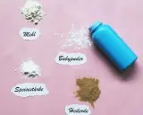 Ein blauer Behälter mit Babypuder liegt neben kleinen Häufchen von Mehl, Speisestärke und Heilerde. Babypuder als Trockenshampoo ist eine schnelle Lösung.