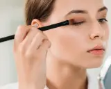 Eine Frau schminkt sich mit einem Pinsel Lidschatten auf, um strahlende Augen zu kreieren. Sie blickt in einen Spiegel.