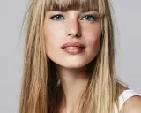 Blonde Frau mit auffälligem Buzz Cut und blauen Augen, die direkt in die Kamera blickt.
