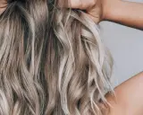 Frau mit blonden, welligen Haaren, die ihre Hände im Nacken hat. Sie zeigt, wie man die Haare pflegt, um sie gesund und glänzend zu halten.