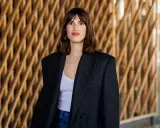Frau mit französischem Crop-Haarschnitt, trägt einen Oversized-Blazer und Jeans vor einer Holzwand.