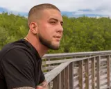 Junger Mann mit Buzzcut und Bart lehnt an einem Holzgeländer, blickt nachdenklich in die Ferne.
