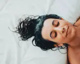Eine Frau mit dunklen, nassen Haaren schläft auf einem weißen Bett. Sie liegt auf dem Rücken, die Augen geschlossen, und ihr Gesicht ist entspannt.