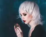 Stylische Dame mit weißem Wolfcut, dunklem Lidschatten und roten Lippen hält eine Brille.