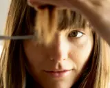 Frau schneidet sich selbst die curtain bangs. Sie hält eine Haarsträhne hoch und setzt die Schere an.