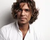 Ein Mann mit braunen, welligen Haaren und Mittelscheitel trägt ein weißes Hemd. Sein Blick ist nachdenklich.