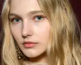 Blonde Haare umrahmen ein Gesicht mit blauen Augen und dezentem Make-up. Ihre hooded eyes wirken geheimnisvoll.