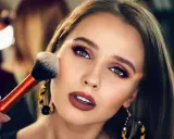 Ein glamouröses Abend Make-up wird aufgetragen. Eine Hand hält einen Pinsel, der über die Wange einer Frau mit dramatischem Lidschatten und Lippenstift gleitet.