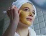 Junge Frau mit Handtuch auf dem Kopf trägt eine Gesichtsmaske auf. Die Kurkuma Wirkung auf die Haut ist bekannt für ihre entzündungshemmenden Eigenschaften.