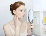 Junge Frau untersucht mit Finger und Spiegel einen Mitesser, der immer wieder an gleicher Stelle kommt.