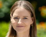 Frau mit blauen Augen und braunem Haar, die lächelt. Ein dezenter Ansatz von grauen Haaren wird durch die Frisur kaschiert.