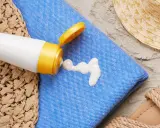 Sonnencreme-Flecken auf dem blauen Handtuch. Ein weißer Klecks Sonnencreme tropft aus einer Tube. Strandutensilien wie Hut und Sandalen liegen daneben.