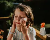 Kind mit geschlossenen Augen trägt Sonnencreme auf. Eine Flasche abgelaufene Sonnencreme steht daneben.