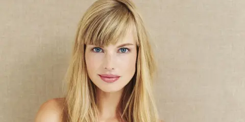 Blonde Frau mit blauen Augen und einem lässigen Pony, der aussieht, als hätte sie ihn selber geschnitten.