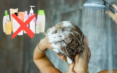 Frau wäscht sich die Haare unter der Dusche, Schaum im Haar. Eine rote durchgestrichene Gruppe von Shampoo-Flaschen deutet auf haare waschen ohne shampoo hin.