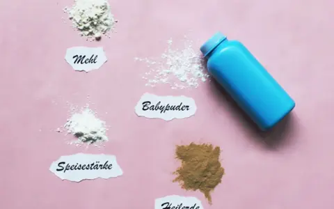 Ein blauer Behälter mit Babypuder liegt neben kleinen Häufchen von Mehl, Speisestärke und Heilerde. Babypuder als Trockenshampoo ist eine schnelle Lösung.