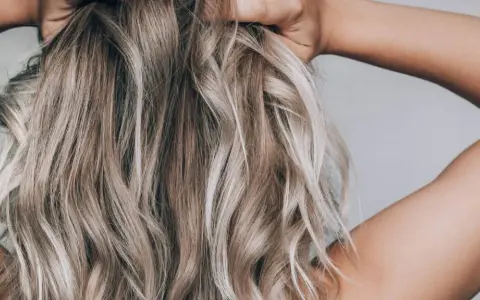Frau mit blonden, welligen Haaren, die ihre Hände im Nacken hat. Sie zeigt, wie man die Haare pflegt, um sie gesund und glänzend zu halten.