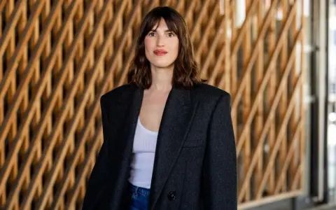Frau mit französischem Crop-Haarschnitt, trägt einen Oversized-Blazer und Jeans vor einer Holzwand.