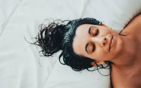 Eine Frau mit dunklen, nassen Haaren schläft auf einem weißen Bett. Sie liegt auf dem Rücken, die Augen geschlossen, und ihr Gesicht ist entspannt.