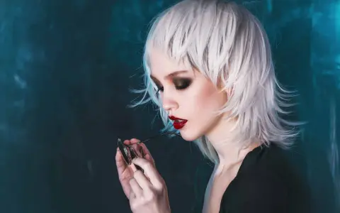 Stylische Dame mit weißem Wolfcut, dunklem Lidschatten und roten Lippen hält eine Brille.