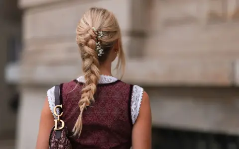Blonde Zopf-Frisur mit Blumen-Akzenten, perfekt für Oktoberfest-Frisuren. Ein Dirndl und eine Dior-Tasche runden den Look ab.