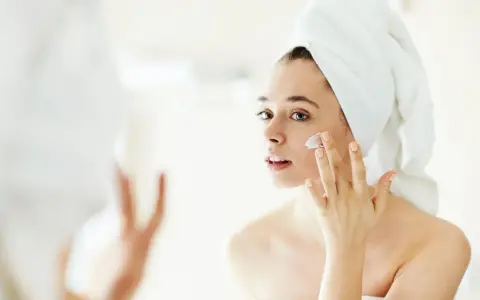 Frau mit Handtuch auf dem Kopf trägt Creme auf, um trockene Haut im Winter zu pflegen.