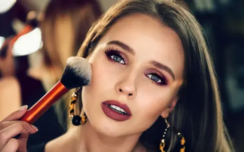 Ein glamouröses Abend Make-up wird aufgetragen. Eine Hand hält einen Pinsel, der über die Wange einer Frau mit dramatischem Lidschatten und Lippenstift gleitet.