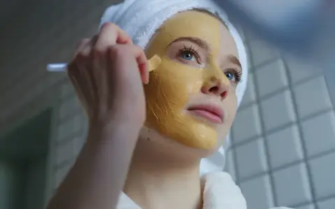 Junge Frau mit Handtuch auf dem Kopf trägt eine Gesichtsmaske auf. Die Kurkuma Wirkung auf die Haut ist bekannt für ihre entzündungshemmenden Eigenschaften.