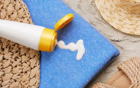 Sonnencreme-Flecken auf dem blauen Handtuch. Ein weißer Klecks Sonnencreme tropft aus einer Tube. Strandutensilien wie Hut und Sandalen liegen daneben.