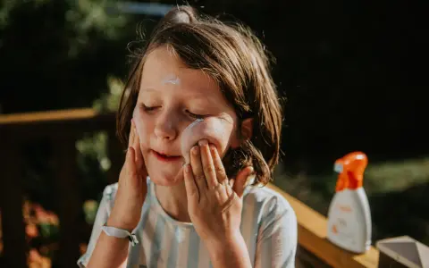 Kind mit geschlossenen Augen trägt Sonnencreme auf. Eine Flasche abgelaufene Sonnencreme steht daneben.