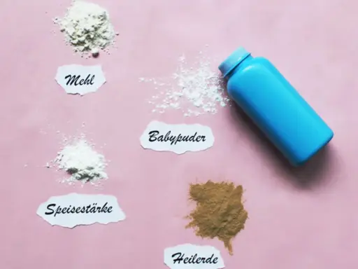 Ein blauer Behälter mit Babypuder liegt neben kleinen Häufchen von Mehl, Speisestärke und Heilerde. Babypuder als Trockenshampoo ist eine schnelle Lösung.