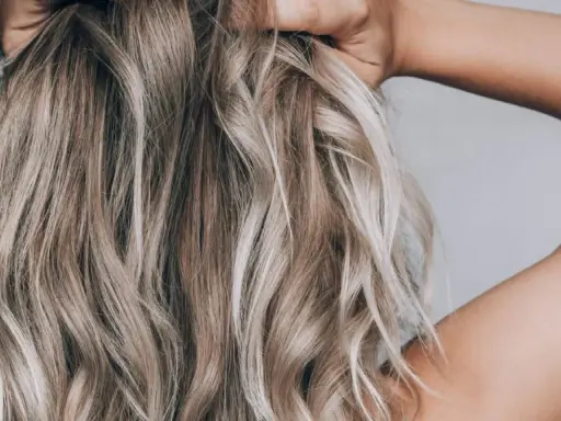 Frau mit blonden, welligen Haaren, die ihre Hände im Nacken hat. Sie zeigt, wie man die Haare pflegt, um sie gesund und glänzend zu halten.