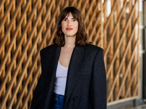 Frau mit französischem Crop-Haarschnitt, trägt einen Oversized-Blazer und Jeans vor einer Holzwand.