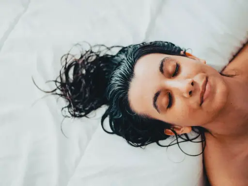 Eine Frau mit dunklen, nassen Haaren schläft auf einem weißen Bett. Sie liegt auf dem Rücken, die Augen geschlossen, und ihr Gesicht ist entspannt.