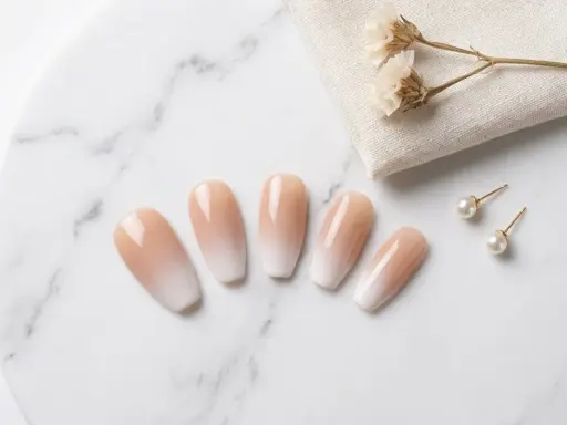 Fünf elegante Ombre-Nägel mit sanftem Farbverlauf von Nude zu Weiß, daneben Perlenohrstecker und getrocknete Blumen.