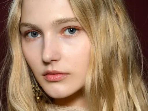 Blonde Haare umrahmen ein Gesicht mit blauen Augen und dezentem Make-up. Ihre hooded eyes wirken geheimnisvoll.