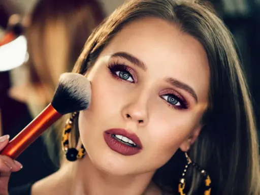 Ein glamouröses Abend Make-up wird aufgetragen. Eine Hand hält einen Pinsel, der über die Wange einer Frau mit dramatischem Lidschatten und Lippenstift gleitet.