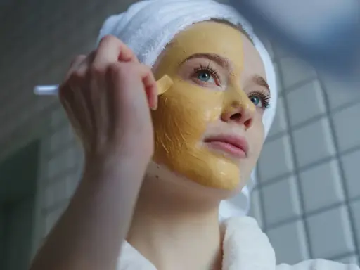 Junge Frau mit Handtuch auf dem Kopf trägt eine Gesichtsmaske auf. Die Kurkuma Wirkung auf die Haut ist bekannt für ihre entzündungshemmenden Eigenschaften.